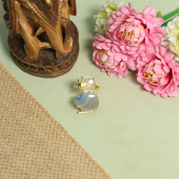 A lifestyle close-up of a woman wearing a large gold geometric ring by Belsi's Jewelry featuring a banded blue lace agate and a faceted clear crystal quartz.