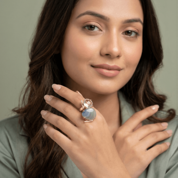 A lifestyle close-up of a woman wearing a large gold geometric ring by Belsi's Jewelry featuring a banded blue lace agate and a faceted clear crystal quartz.