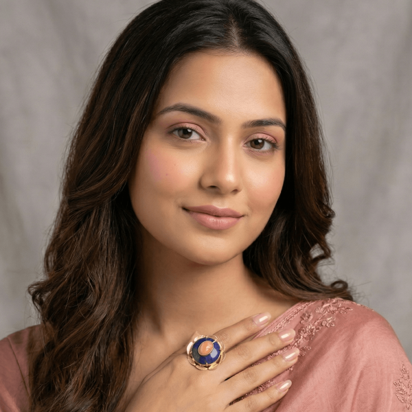 A lifestyle shot of a woman in a mauve embroidered saree wearing a large round gold floral ring with a tiered blue lapis lazuli and peach sunstone center.