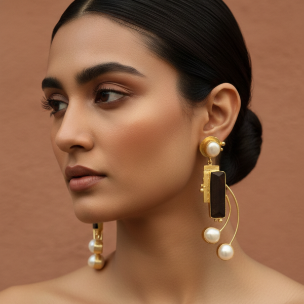 Model wearing long handcrafted gold-tone statement earrings featuring a dark rectangular centerpiece and suspended baroque pearls.