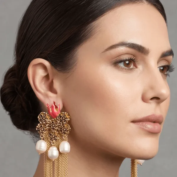 Close-up studio profile portrait of a model showcasing Belsi's Jewelry Flower Power collection long gold tassel earrings, designed with a textured floral cluster stud, bright coral pink enamel accents, and hanging freshwater pearls with fluid gold chain fringes.