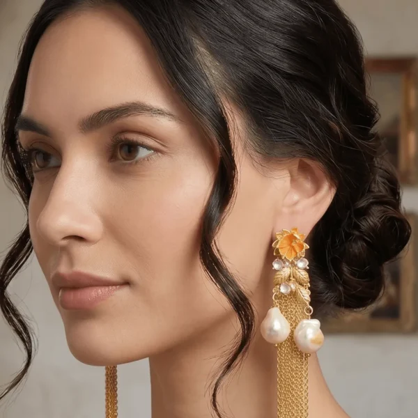 Studio profile portrait of a model showcasing Belsi's Jewelry Flower Power collection long statement earrings, featuring a warm ochre orange enamel top flower, a gold leaf vine drop structure accented with clear stones, and two hanging baroque pearls with ultra-fine gold chain fringes.