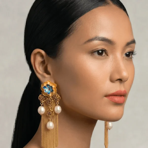 Close-up profile portrait of a model showcasing Belsi's Jewelry Flower Power collection long statement earrings, featuring a large tiered gold flower stud with deep indigo and sky blue enamel petals, a cluster drop of smaller filigree roses, multiple hanging round freshwater pearls, and long fluid gold chain fringe tassels.