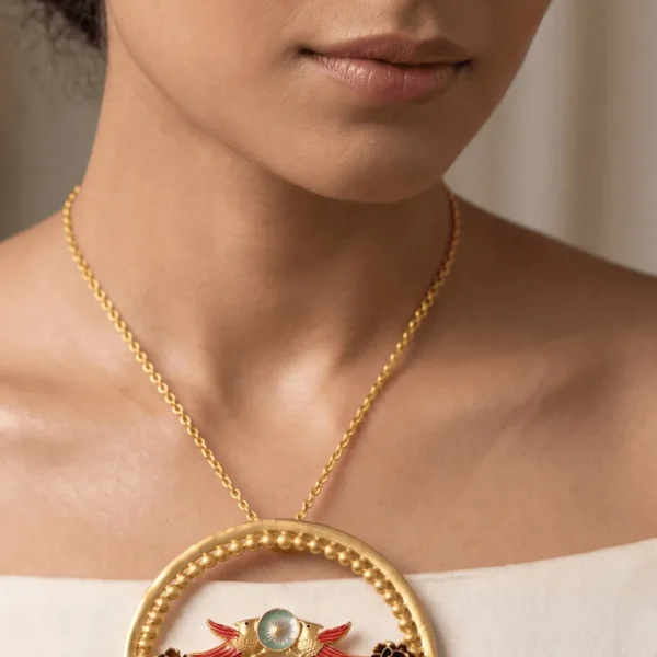 High-fashion editorial shot of a model wearing an oversized circular matte-finish gold statement pendant with handcrafted bird motifs, lime green enamel detailing, and a prominent pink teardrop gemstone.