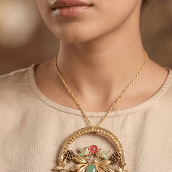 Studio portrait of a model wearing an oversized circular matte-gold statement pendant necklace featuring mint green artisan birds, crimson enamel rosettes, and a teardrop mint gemstone.