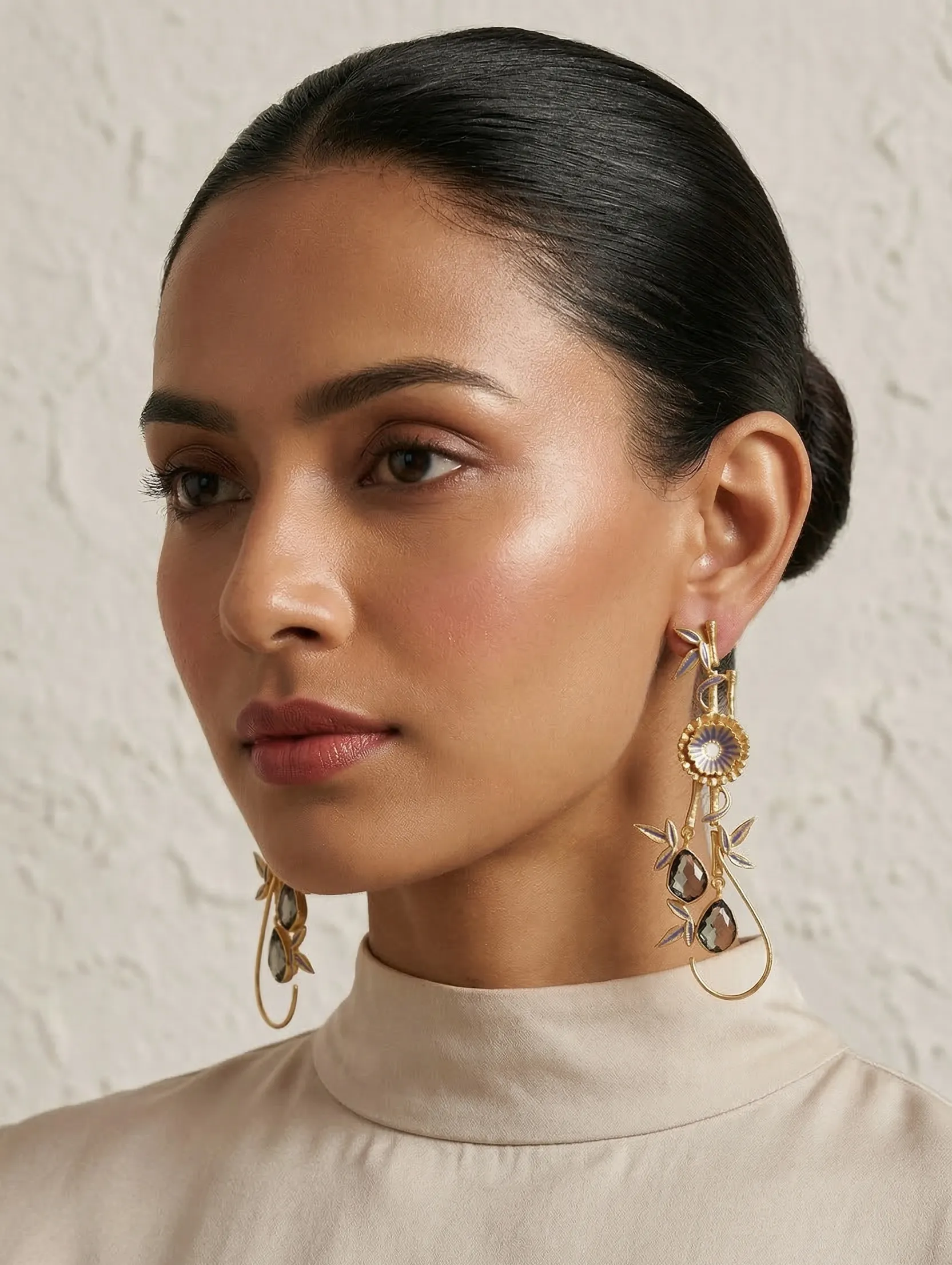 Aura Enamelled Bamboo & Floral Drop Earrings in Gold-Tone with Mauve & Smoky Gemstones 1 Close-up of a model wearing long gold-tone statement drop earrings featuring muted mauve and lavender enamel bamboo accents, a central floral motif, and teardrop-shaped smoky grey gemstones suspended on a wire loop.