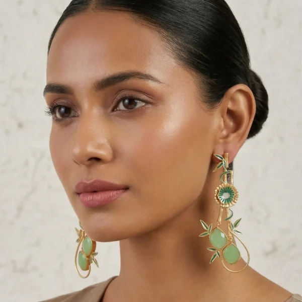 Model wearing long gold-tone statement drop earrings featuring jade-green enamel bamboo accents, a central floral motif, and natural green oval gemstones on a delicate wire loop.