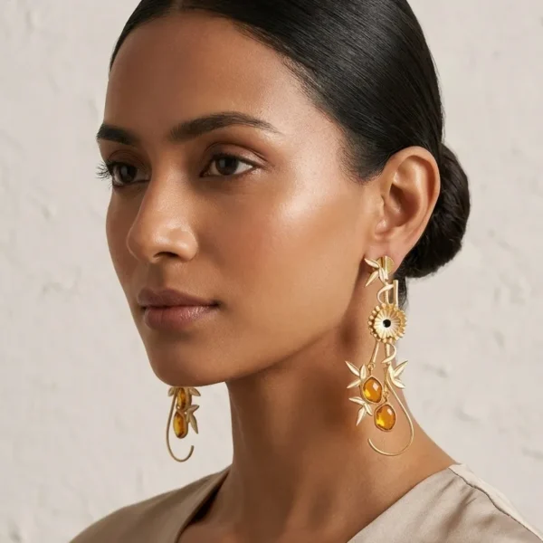 Model wearing long gold-tone statement drop earrings featuring polished gold bamboo accents, a central floral motif, and warm honey-amber oval gemstones on a delicate wire loop.