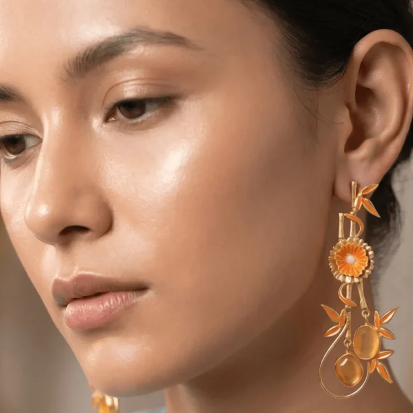 Close-up of a model wearing long gold-tone statement drop earrings featuring vibrant tangerine-orange enamel bamboo accents, a central floral motif, and two matching translucent orange oval gemstones suspended on a wire loop.