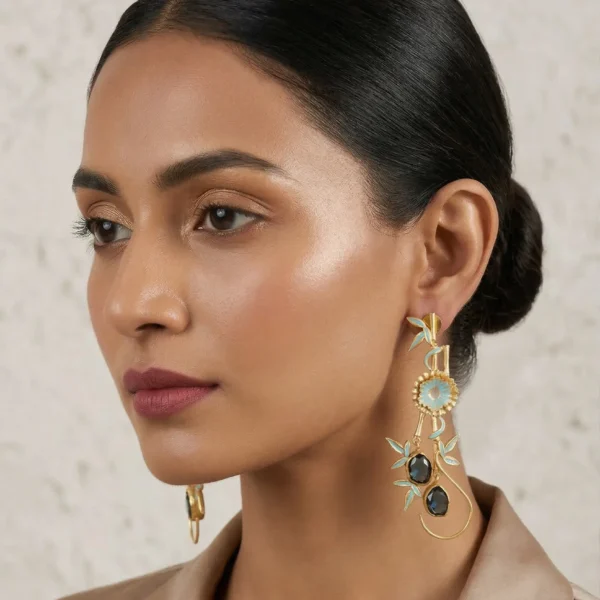 Close-up of a woman wearing a long, gold-tone statement drop earring featuring light blue enamel bamboo leaves, a central floral motif, and two dark oval gemstones suspended on a curved wire loop.