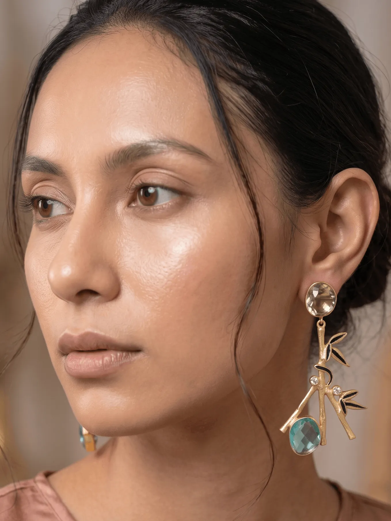 Zen Enamelled Bamboo Branch Drop Earrings with Teal & Champagne Gemstones 1 Close-up of a model wearing gold-tone statement earrings shaped like a textured bamboo branch, featuring black enamel leaf accents, clear crystal detailing, a champagne round top stone, and a bright teal blue teardrop gemstone.