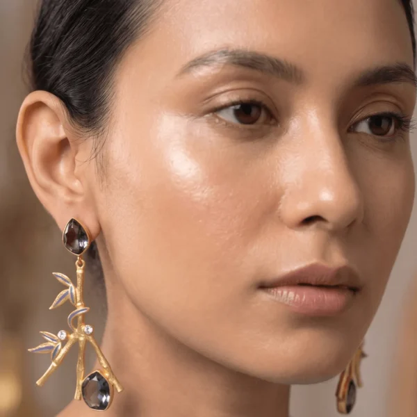 Close-up of a model wearing a gold-tone statement earring shaped like a textured bamboo branch, featuring blue enamel leaf detailing, tiny sparkling cubic zirconia accents, and smoky grey teardrop gemstones.