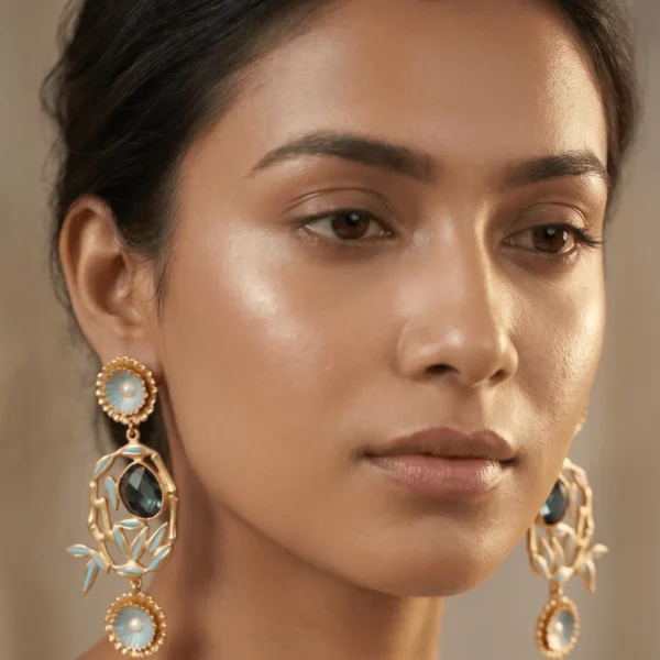Close-up of a model wearing large, ornate gold-tone chandelier statement earrings featuring dual powder blue enamel floral medallions, a matching blue enameled central bamboo frame, and a dark faceted oval centerpiece gemstone.