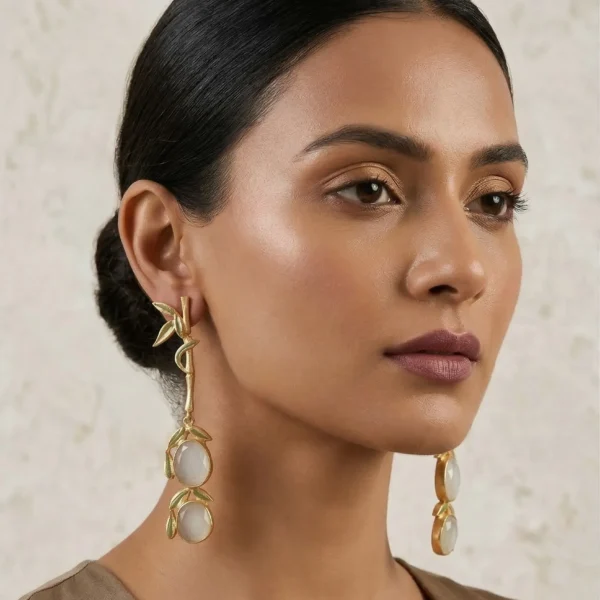 Close-up of a model wearing long gold-tone linear statement earrings featuring a single textured bamboo stalk with sage green enamel leaves, leading down to two stacked milky white oval gemstones framed in matching enamel.