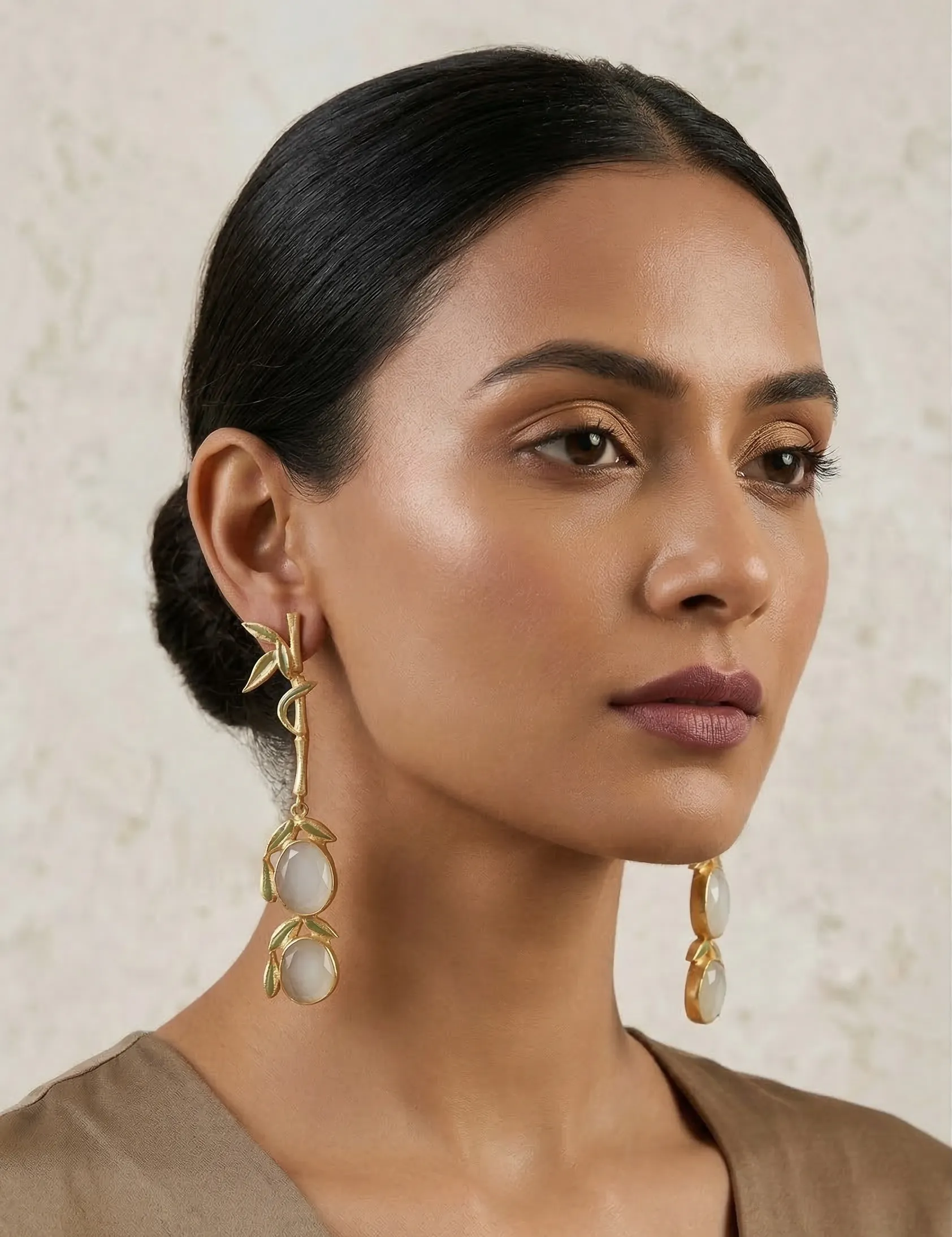 Majestic Enamelled Bamboo Drop Earrings in Gold-Tone with Milky White Gemstones 1 Close-up of a model wearing long gold-tone linear statement earrings featuring a single textured bamboo stalk with sage green enamel leaves, leading down to two stacked milky white oval gemstones framed in matching enamel.