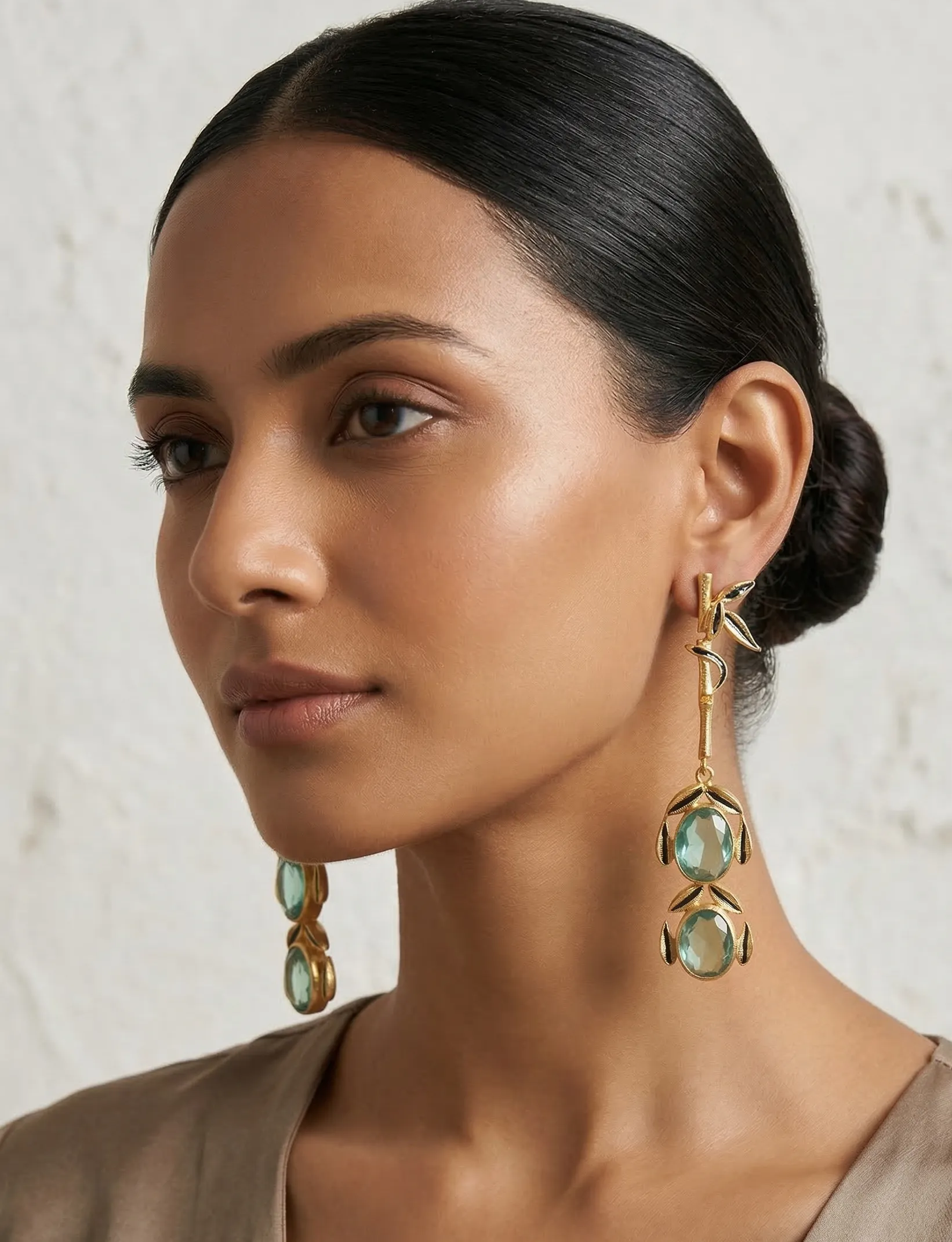Majestic Enamelled Bamboo Drop Earrings in Gold-Tone with Seafoam Green Gemstones 1 Close-up of a model wearing long gold-tone linear statement earrings featuring a single textured bamboo stalk with black enamel leaves, leading down to two stacked faceted seafoam green oval gemstones framed in matching enamel.