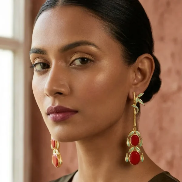 Close-up of a model wearing long gold-tone linear statement earrings featuring a single textured bamboo stalk with light green enamel leaves, leading down to two stacked ruby-red oval gemstones framed in matching enamel.