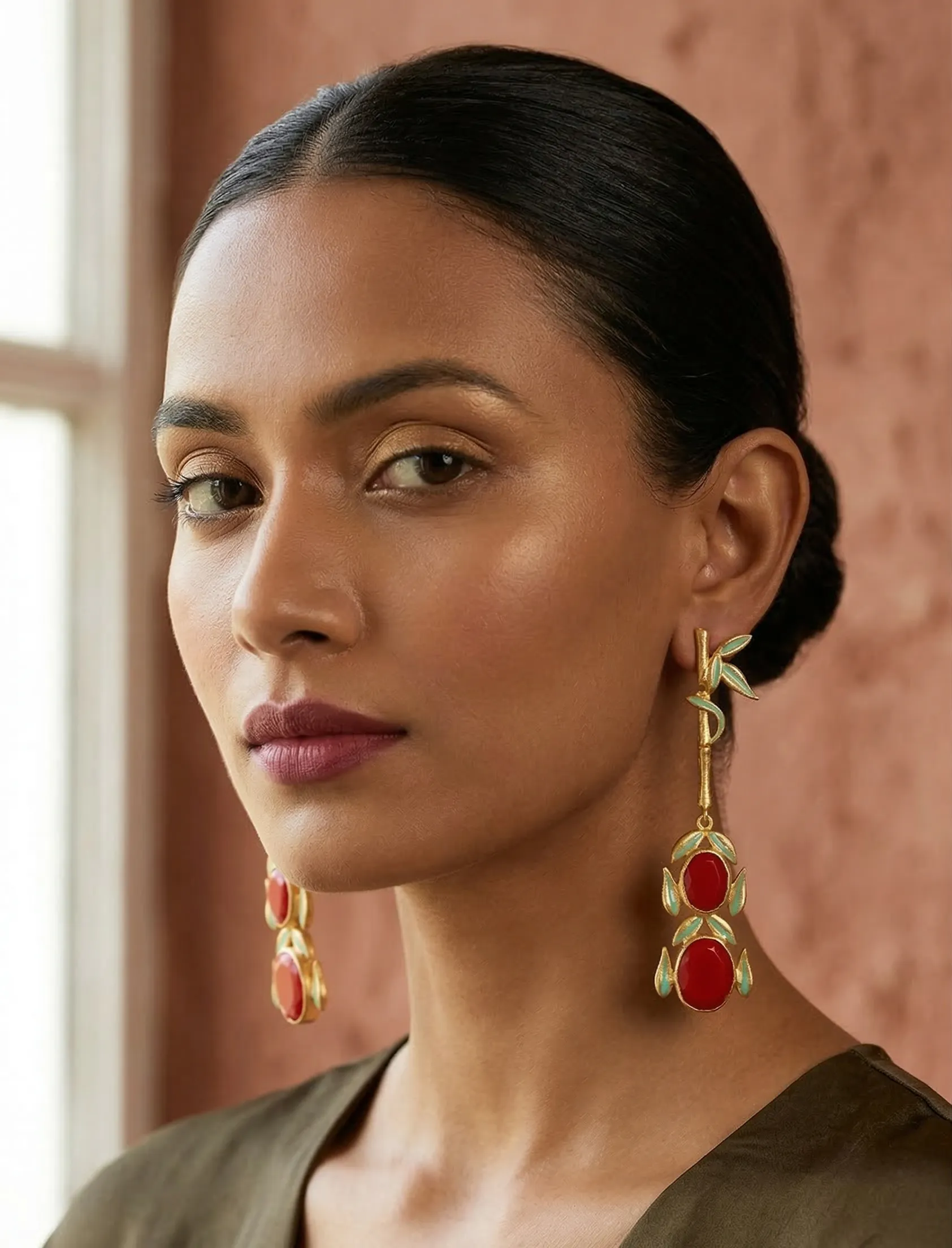 Majestic Enamelled Bamboo Drop Earrings in Gold-Tone with Ruby Red Gemstones 1 Close-up of a model wearing long gold-tone linear statement earrings featuring a single textured bamboo stalk with light green enamel leaves, leading down to two stacked ruby-red oval gemstones framed in matching enamel.