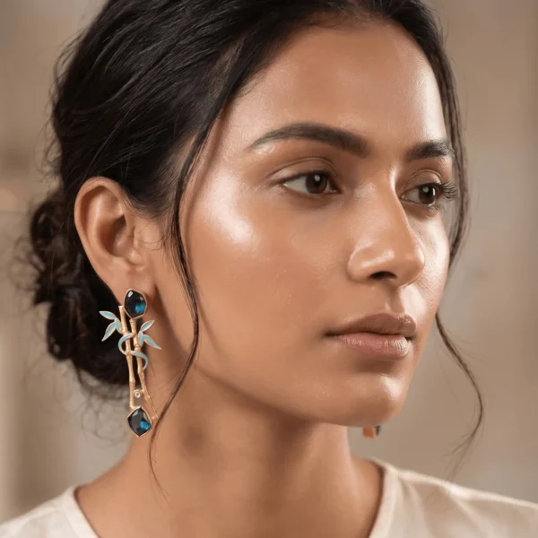 Close-up of a model wearing gold-tone statement earrings shaped like a textured bamboo branch, featuring soft powder blue enamel leaf details, a crystal node accent, and two dark faceted deep teal blue gemstones.