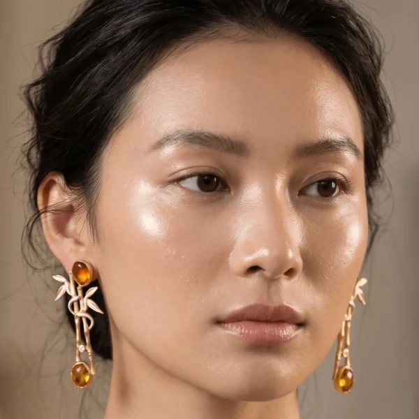 Close-up of a model wearing gold-tone statement earrings shaped like a textured bamboo branch, featuring soft ivory-white enamel leaves, a crystal node accent, and two warm translucent amber-toned gemstones.