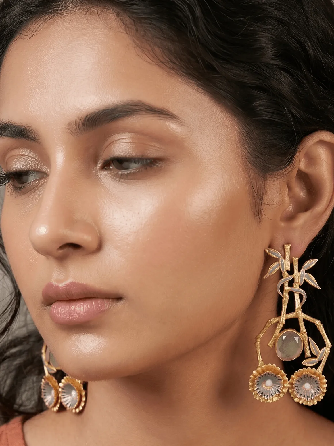 Imperial Enamelled Bamboo Chandelier Earrings in Gold-Tone with Slate Grey Accents & Milky Stone 1 Close-up of a model wearing large, ornate gold-tone chandelier statement earrings featuring slate grey and off-white enamel accents on a structural bamboo framework, a central faceted translucent milky grey gemstone, and dual fluted floral wheels at the base.