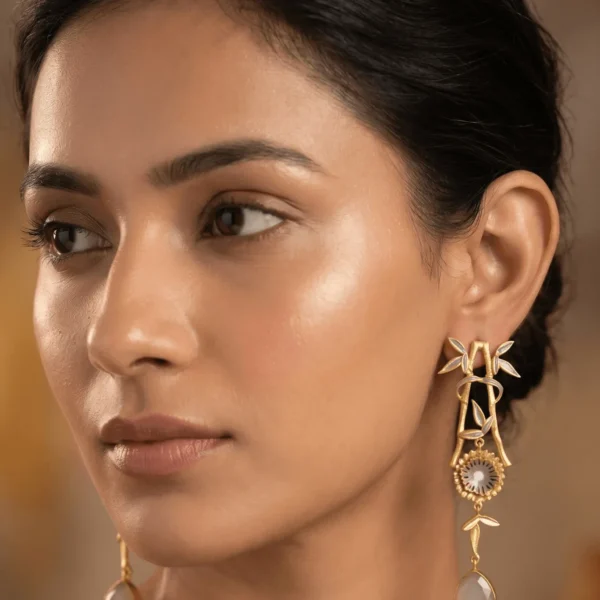 Close-up of a model wearing long gold-tone linear statement earrings featuring muted slate grey and off-white enamel accents on an architectural bamboo frame, a central fluted floral medallion, and a translucent milky grey teardrop gemstone dangling at the base.