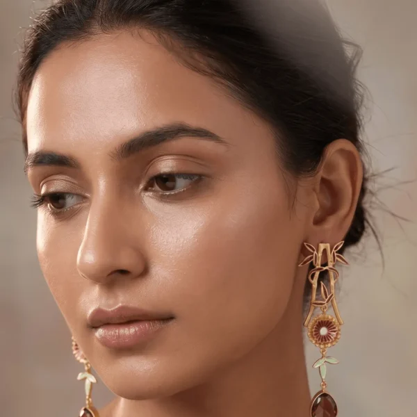 Close-up of a model wearing long gold-tone linear statement earrings featuring deep maroon and off-white enamel accents on an architectural bamboo frame, a central fluted floral medallion, and a faceted amber brown teardrop gemstone dangling at the base.