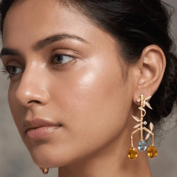 ose-up of a model wearing gold-tone statement earrings designed like a branched bamboo stalk, featuring pale peach enamel leaves, sparkling diamond-like node accents, two teardrop amber gemstones flanking the outer branches, and a central oval sky blue gemstone.