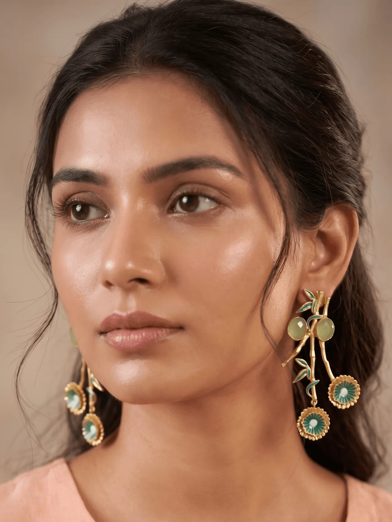 Blossom Enamelled Bamboo Branch Chandelier Earrings with Sage Green Gemstones & Seafoam Accents 1 Close-up of a model wearing large gold-tone branched statement earrings resembling a bamboo stalk with rich seafoam teal-green enamel leaves, two smooth round milky sage green gemstones, and twin fluted floral medallions hanging at the base.