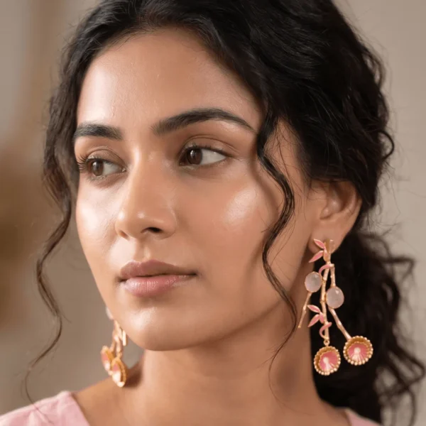 Close-up of a model wearing large gold-tone branched statement earrings resembling a bamboo stalk with bright pink enamel leaves, two smooth round translucent rose quartz gemstones, and twin fluted floral medallions hanging at the base.