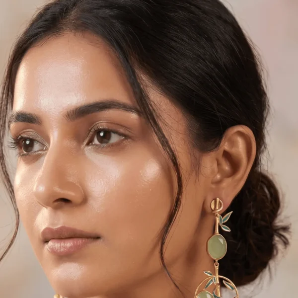 Close-up of a model wearing long gold-tone statement earrings featuring a linear bamboo stalk with seafoam teal-green enamel leaves, a central oval milky sage green gemstone, and a dangling circular frame encasing an intricate bamboo branch motif and matching smooth sage green stones.