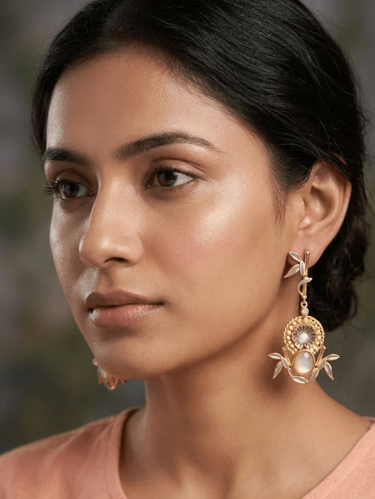 Vintage Enamelled Bamboo Medallion Drop Earrings with Moonstone White Gemstones 1 Close-up of a model wearing gold-tone statement drop earrings featuring a linear bamboo post with soft ivory enamel leaves, a central fluted concentric medallion filled with matching ivory accents, and a nested oval milky pearlescent white gemstone framed by hand-painted botanical foliage.