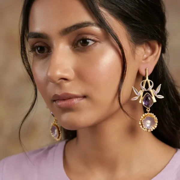 Close-up of a model wearing gold-tone statement drop earrings featuring a linear post, an openwork bamboo frame forming a shield silhouette with soft lavender enamel accents, a central faceted teardrop purple amethyst gemstone, and a tiered fluted orchid medallion dangling at the base.