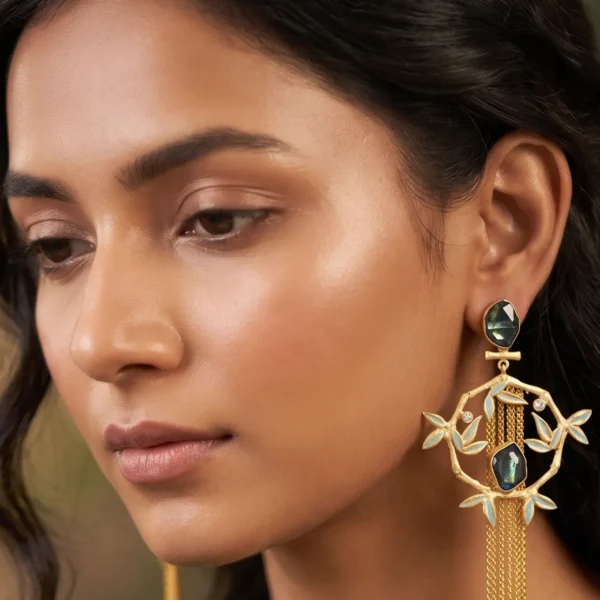 Close-up of a model wearing long gold-tone statement fringe earrings featuring an oval dark faceted teal gemstone stud, a dangling diamond-shaped bamboo frame accented with light powder blue enamel leaves, a central matching dark teal stone, and a long cascading waterfall of fine gold chain tassels.