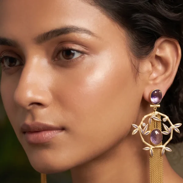 Close-up of a model wearing long gold-tone statement fringe earrings featuring an oval faceted purple amethyst stud, a dangling diamond-shaped bamboo frame accented with soft lavender enamel leaves, a central matching oval purple gemstone, and a long cascading waterfall of fine gold chain tassels.