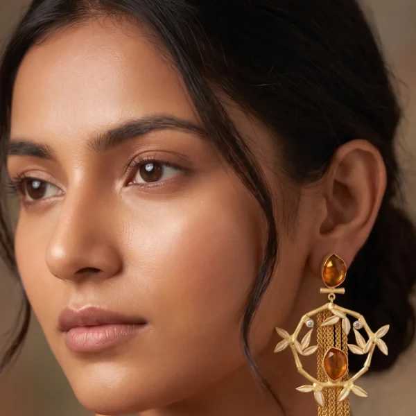 Close-up of a model wearing long gold-tone statement fringe earrings featuring an oval faceted honey amber stud, a dangling diamond-shaped bamboo frame accented with cream enamel leaves, a central matching oval amber gemstone, and a long cascading waterfall of fine gold chain tassels.