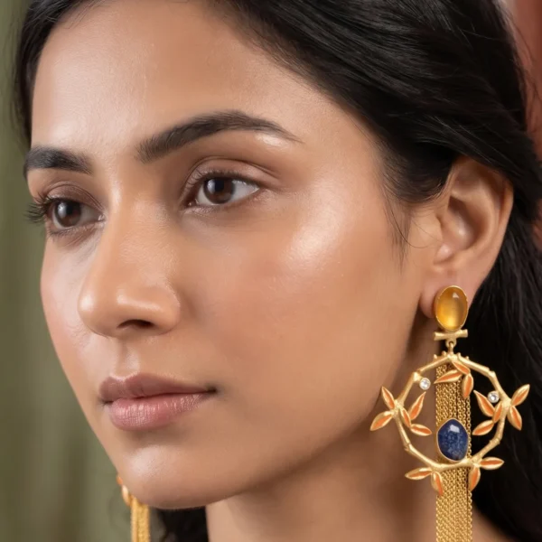 Close-up of a model wearing long gold-tone statement fringe earrings featuring an oval translucent yellowish-brown amber stud stone, a dangling diamond-shaped bamboo frame accented with bright saffron orange enamel leaves, a central oval royal blue lapis-style gemstone, and a long cascading waterfall of fine gold chain tassels.