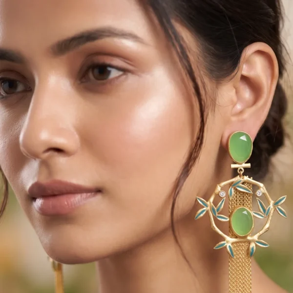Close-up of a model wearing long gold-tone statement fringe earrings featuring an oval milky sage green stud, a dangling diamond-shaped bamboo wreath frame accented with seafoam teal enamel leaves, a central matching oval green gemstone, and a long cascading waterfall of fine gold chain tassels.