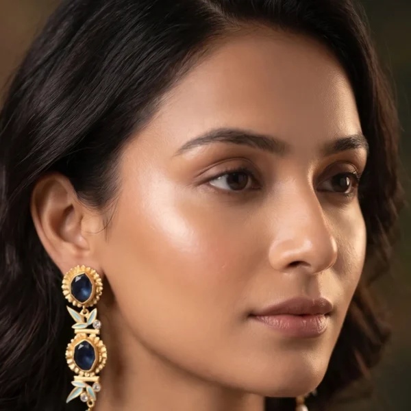 Close-up of a model wearing gold-tone linear statement earrings featuring a tiered arrangement of two fluted concentric medallions set with faceted royal navy blue stones, separated by soft powder blue enamel bamboo leaves and clear crystal nodes, finished with a dangling organic white baroque pearl.