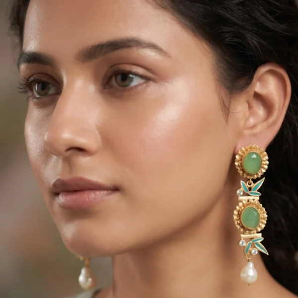 Close-up of a model wearing gold-tone linear statement earrings featuring a tiered arrangement of two fluted concentric medallions set with milky sage green stones, separated by seafoam teal enamel bamboo leaves and clear crystal nodes, finished with a dangling organic white baroque pearl.
