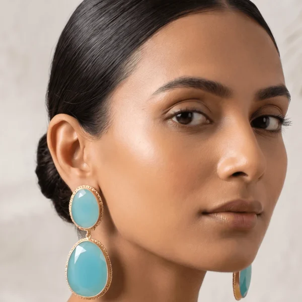 Close-up editorial portrait of a woman with a sleek low bun wearing translucent aqua blue double drop statement earrings with textured gold-toned borders from the Spectrum Luxe Collection.