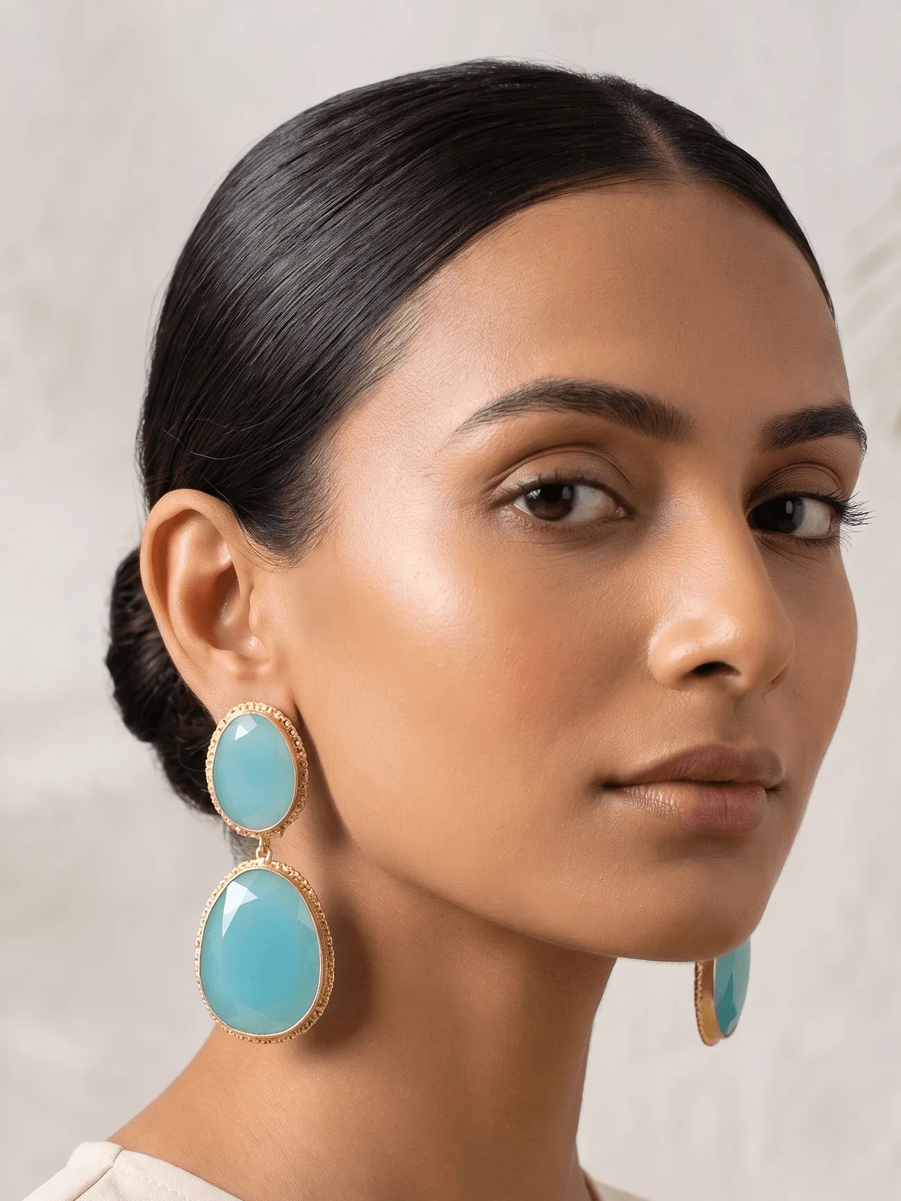 Aqua Blue Textured Gold Dual Drop Earrings 1 Close-up editorial portrait of a woman with a sleek low bun wearing translucent aqua blue double drop statement earrings with textured gold-toned borders from the Spectrum Luxe Collection.